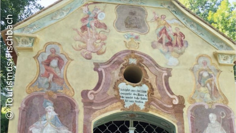Fassade einer Kapelle mit religiösen Fresken und Inschrift.