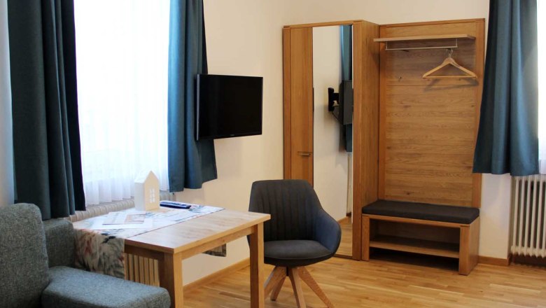 Modernes G&auml;stezimmer mit Tisch, Stuhl, Kleiderschrank, Sofa, Fernseher und blauen Vorh&auml;ngen.