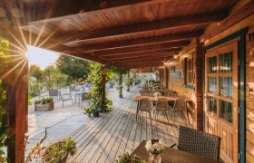 Holzterrasse mit Tischen und St&uuml;hlen, umgeben von Pflanzen, bei Sonnenuntergang.