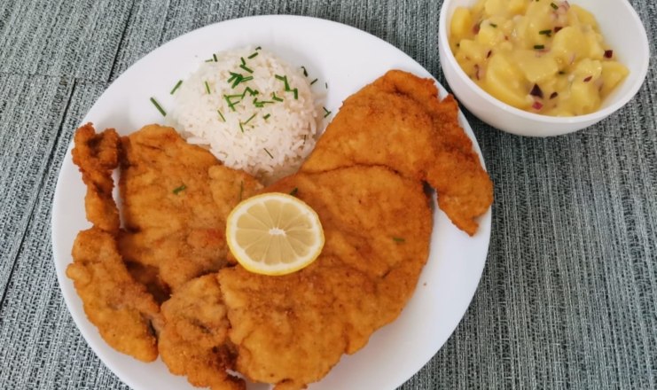 Ein Teller mit paniertem Schnitzel, Reis und einer Zitronenscheibe, daneben eine Sch&uuml;ssel Kartoffelsalat.