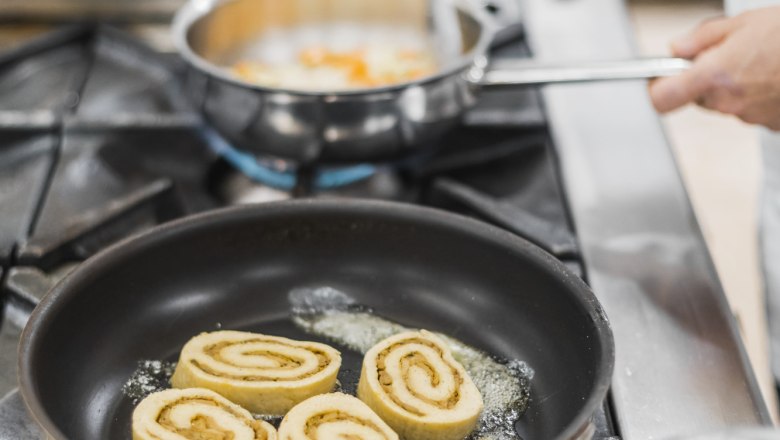 Egy serpenyő három porcini gombás rolóval a tűzhelyen, mellette egy kéz, amely egy másik serpenyőt tart.
