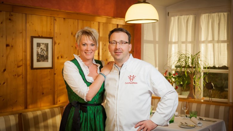 Forsthaus Fischbach, © Werner Krug Ein Mann und eine Frau stehen in einem gemütlichen Restaurant. Die Frau trägt ein Dirndl, der Mann eine Kochjacke mit der Aufschrift 'Zum Forsthaus'.