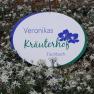 Wei&szlig;es Schild mit lila und gr&uuml;ner Aufschrift "Veronikas Kr&auml;uterhof Fischback" in einem Feld mit Edelwei&szlig;blumen.