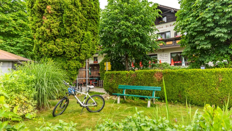 Ein Fahrrad steht neben einer Bank im Garten eines Gasthofs, umgeben von Bäumen und Sträuchern.