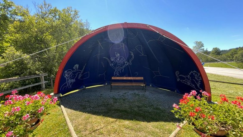 Halbrunde Struktur mit Sternbildern und Bank, umgeben von Blumen und Bäumen.