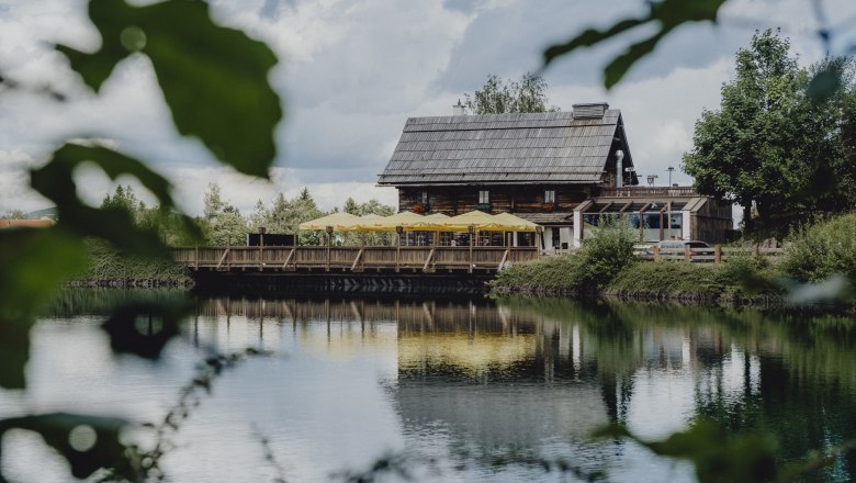 Ein rustikales Holzhaus mit gelben Sonnenschirmen spiegelt sich in einem ruhigen See, umgeben von gr&uuml;ner Natur.
