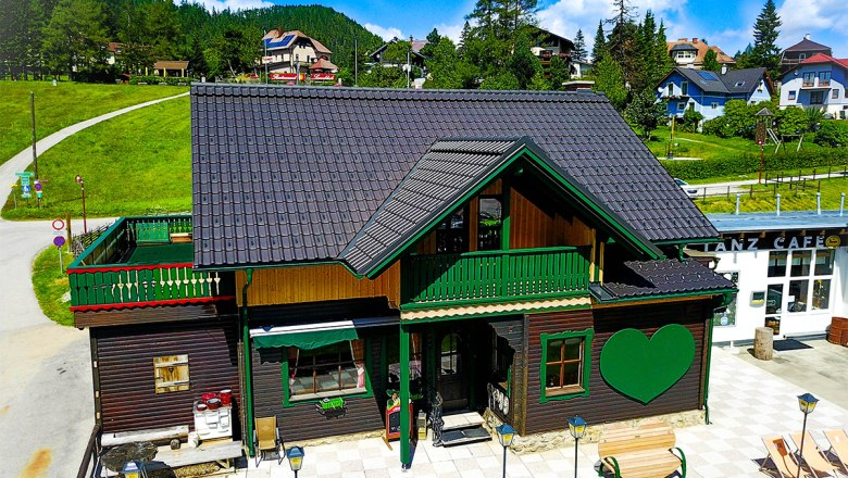 Ein traditionelles Holzhaus mit grünem Herzmotiv und Terrasse im Sommer, umgeben von grünen Hügeln und blauem Himmel.