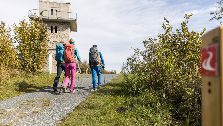 Drei Wanderer mit Rucksäcken gehen auf einem Weg zu einem Steinturm.