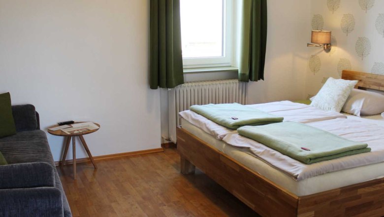 Schlafzimmer mit Doppelbett, Fenster mit grünen Vorhängen, Sofa, Tisch, Tapete und einer Wandlampe.