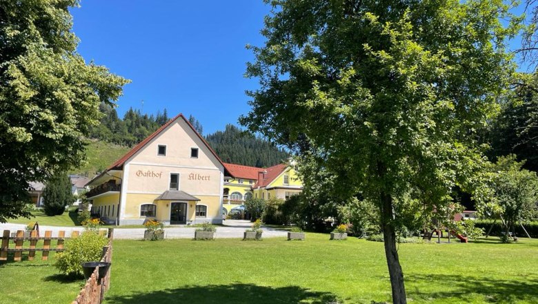 Ein Gasthof mit gelber Fassade und rotem Dach, umgeben von gr&uuml;ner Wiese und B&auml;umen.