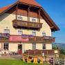 Ein traditionelles Gasthaus mit Holzbalkonen und Blumen, umgeben von gr&uuml;nen H&uuml;geln und blauem Himmel.