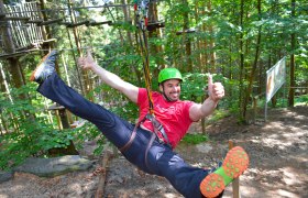 Person mit Helm und Sicherheitsgurt auf einer Seilrutsche im Wald.
