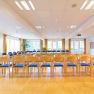 Ein heller Seminarraum mit Stuhlreihen, gro&szlig;en Fenstern und einem Projektor an der Decke.