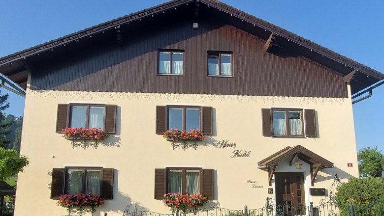 Ein traditionelles zweist&ouml;ckiges Haus mit Holzverkleidung und Blumenk&auml;sten vor den Fenstern.