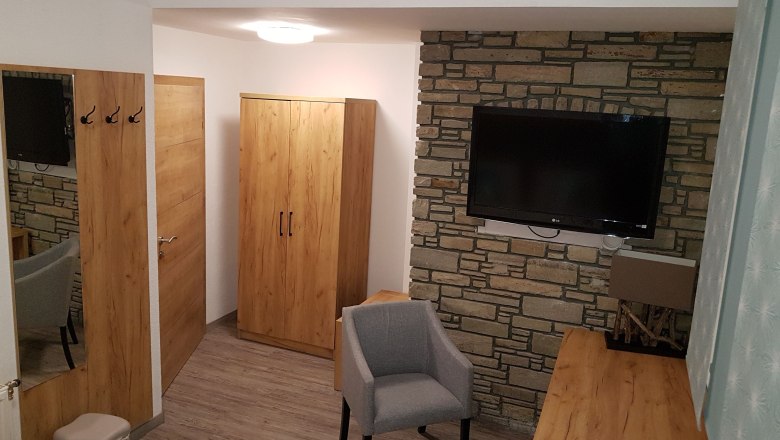 Hotelzimmer mit Holzm&ouml;beln, grauem Sessel und Flachbildfernseher an einer Steinwand.