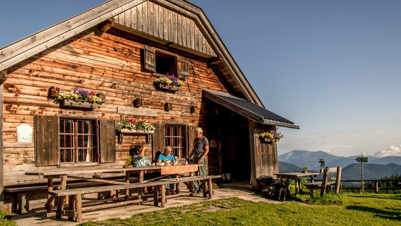 Holzh&uuml;tte in den Bergen mit Menschen am Tisch, blauer Himmel.