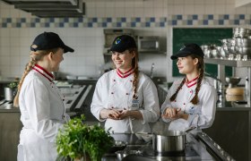 Drei junge Frauen in Kochuniformen und schwarzen Kappen stehen in einer K&uuml;che und unterhalten sich.