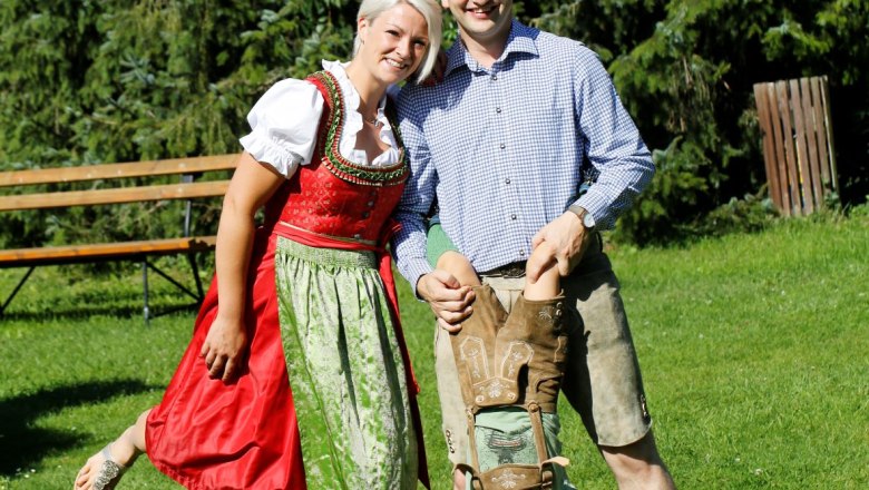 Ein Paar in traditioneller Tracht mit einem Kind, das kopfüber gehalten wird, auf einer Wiese vor Bäumen.