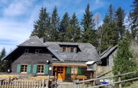 Eine rustikale Berghütte mit Holzzaun und Tischen im Freien, umgeben von hohen Tannenbäumen unter blauem Himmel.