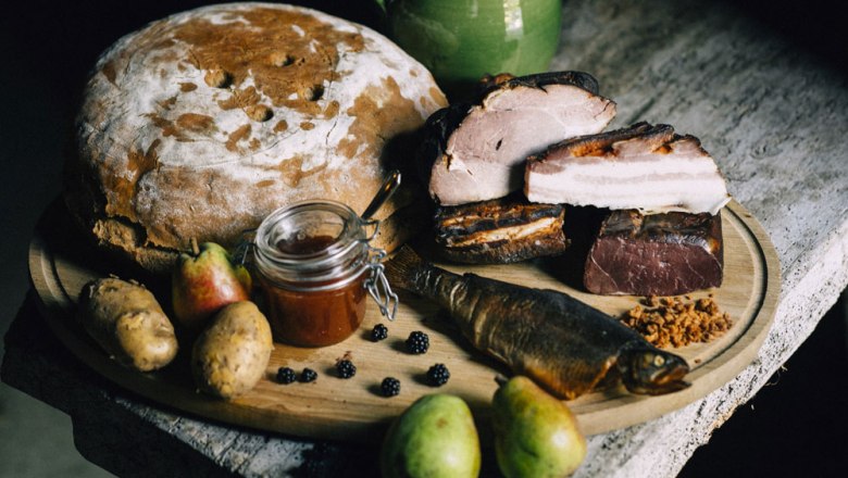 Ein rustikales Arrangement mit Brot, Fleisch, Fisch, Birnen, Kartoffeln und Marmelade auf einem Holztisch.
