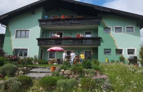 Egy z&ouml;ld h&aacute;z, előtte erk&eacute;lyekkel &eacute;s emberekkel, vir&aacute;gos kerttel k&ouml;r&uuml;lv&eacute;ve.