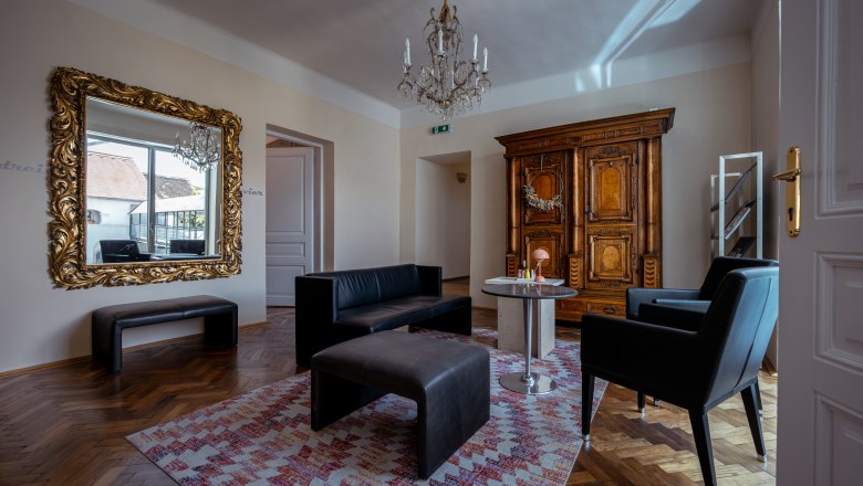 Elegantes Wohnzimmer mit antikem Schrank, großem Spiegel, schwarzen Ledermöbeln und einem Kronleuchter.