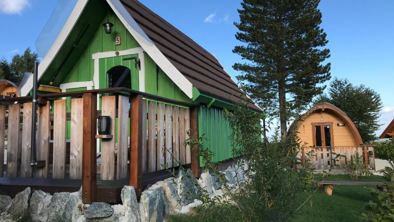 Zwei kleine Holzh&uuml;tten im Glamping-Stil mit gr&uuml;ner und brauner Fassade, umgeben von B&auml;umen und Gras.