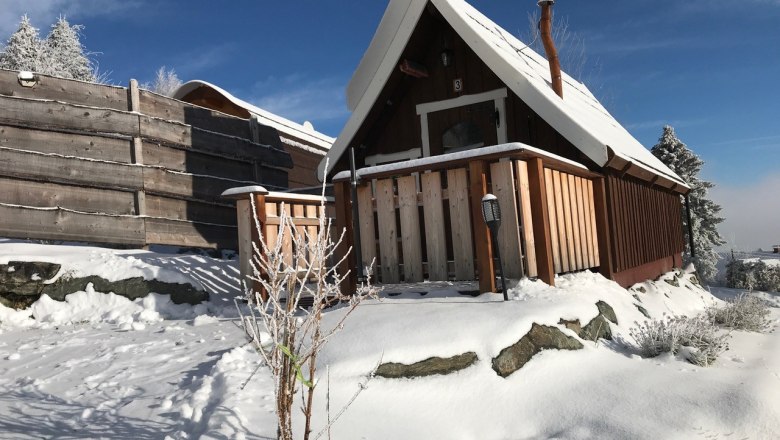 Eine verschneite Holzhütte mit spitzem Dach und Veranda in winterlicher Landschaft.