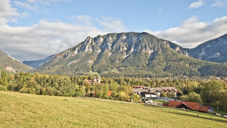 Panoramablick auf eine Berglandschaft mit Wiesen im Vordergrund und einem Dorf im Hintergrund.