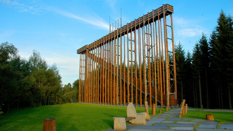 Eine hohe Holzkonstruktion im Freien, umgeben von B&auml;umen und gr&uuml;nem Gras.