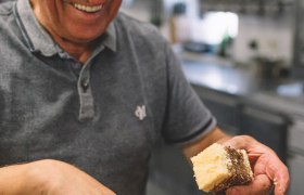 Ein &auml;lterer Mann in einer K&uuml;che h&auml;lt ein St&uuml;ck Kuchen und l&auml;chelt.
