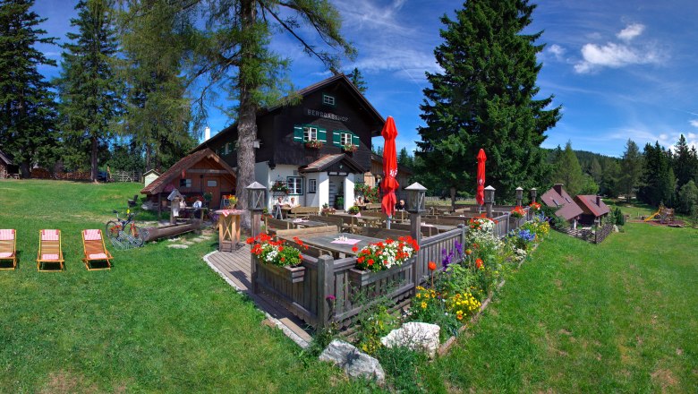 Ein Berggasthof mit Terrasse, Blumen und Liegestühlen auf einer grünen Wiese, umgeben von Bäumen und einem klaren blauen Himmel.