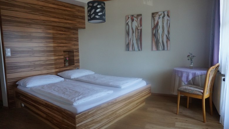 Ein Schlafzimmer mit einem Doppelbett, Holzwandverkleidung, zwei abstrakten Gemälden und einem kleinen Tisch mit Stuhl.