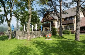 Ein traditionelles Landhaus mit einer mit Holzzaun umrandeten Terrasse, davor eine gr&uuml;ne Wiese,mit hohen B&auml;umen