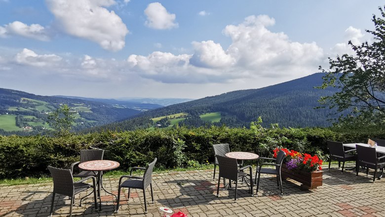 Terrasse mit Tischen und Stühlen, Blick auf grüne Hügel und blauen Himmel mit Wolken.