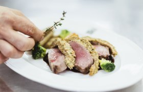 Nahaufnahme eines rosa gebratenen Fleischgerichts mit Kr&auml;uterkruste, Brokkoli und einer Hand, die Thymian hinzuf&uuml;gt.
