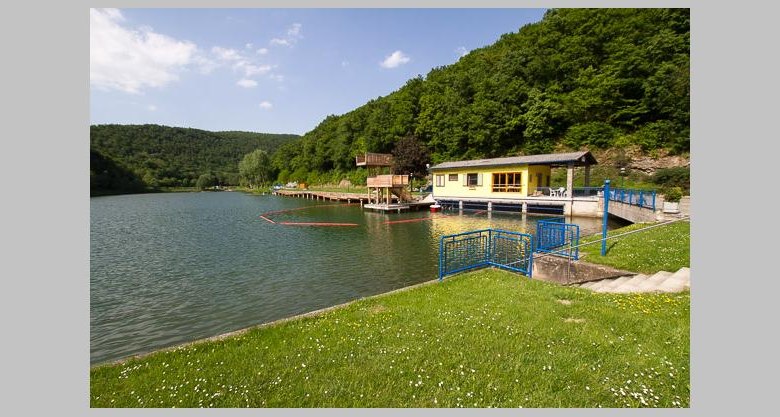 Ein See mit einem gelben Gebäude am Ufer, umgeben von grünen Hügeln und Bäumen.