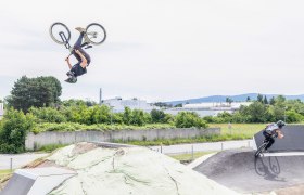 Zwei BMX-Fahrer auf einer Pumptrack-Anlage, einer macht einen R&uuml;ckw&auml;rtssalto, der andere f&auml;hrt eine Kurve.