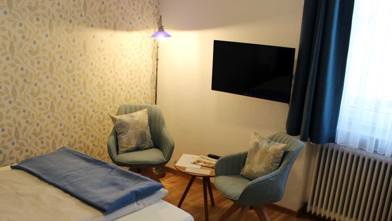 Zimmer mit Bett, Tisch mit zwei St&uuml;hlen, Fernseher, Fenster mit blauen Vorh&auml;ngen und Stehlampe.