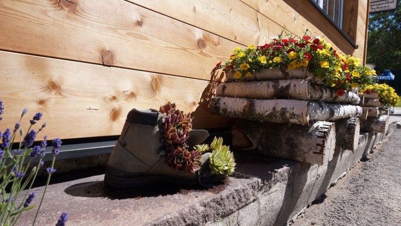 Blumenarrangement mit Wanderschuh und Holzst&auml;mmen vor Holzwand.