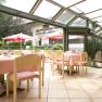 Helles Restaurant mit Glasdach, rosa Tischdecken und Blick auf Garten mit Sonnenschirmen.