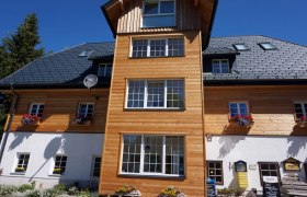 Fassade eines Alpengasthofs mit Holzverkleidung und Blumenkästen unter blauem Himmel.